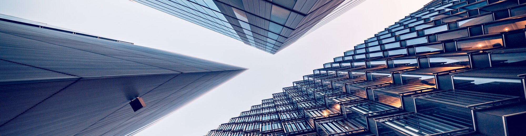 Upward view of modern glass and steel high-rise buildings converging toward the sky, with contrasting facade patterns and glowing window lights.