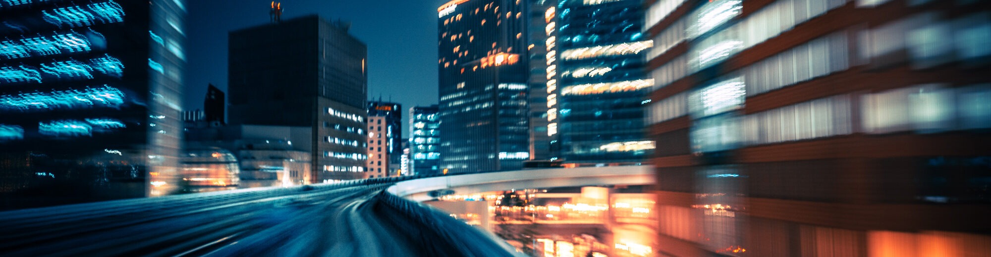 Motion-blurred night-time view along an elevated urban track, with illuminated high-rise buildings streaking past on both sides.