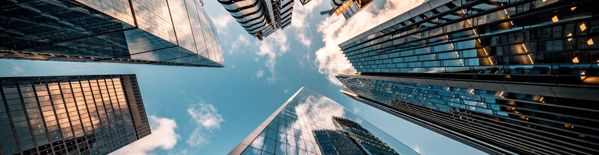 Wide-angle upward view of London skyscrapers, including the Lloyd's building and the Gherkin, converging toward a blue sky with scattered clouds.