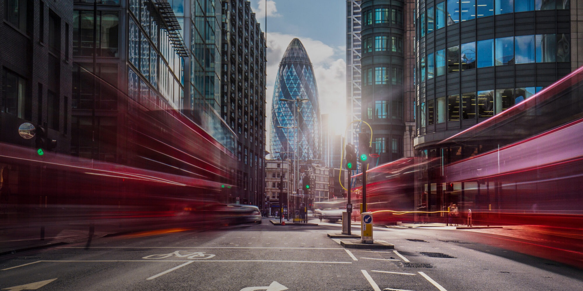 Long-exposure shot of a City of London street with red light trails from passing double-decker buses, the 30 St Mary Axe office building (‚The Gherkin') backlit in the background.